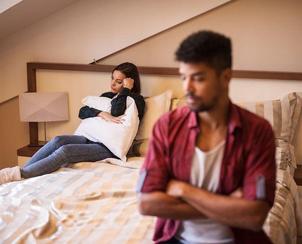 A couple sits on a bed turned away from each other, disgruntled.